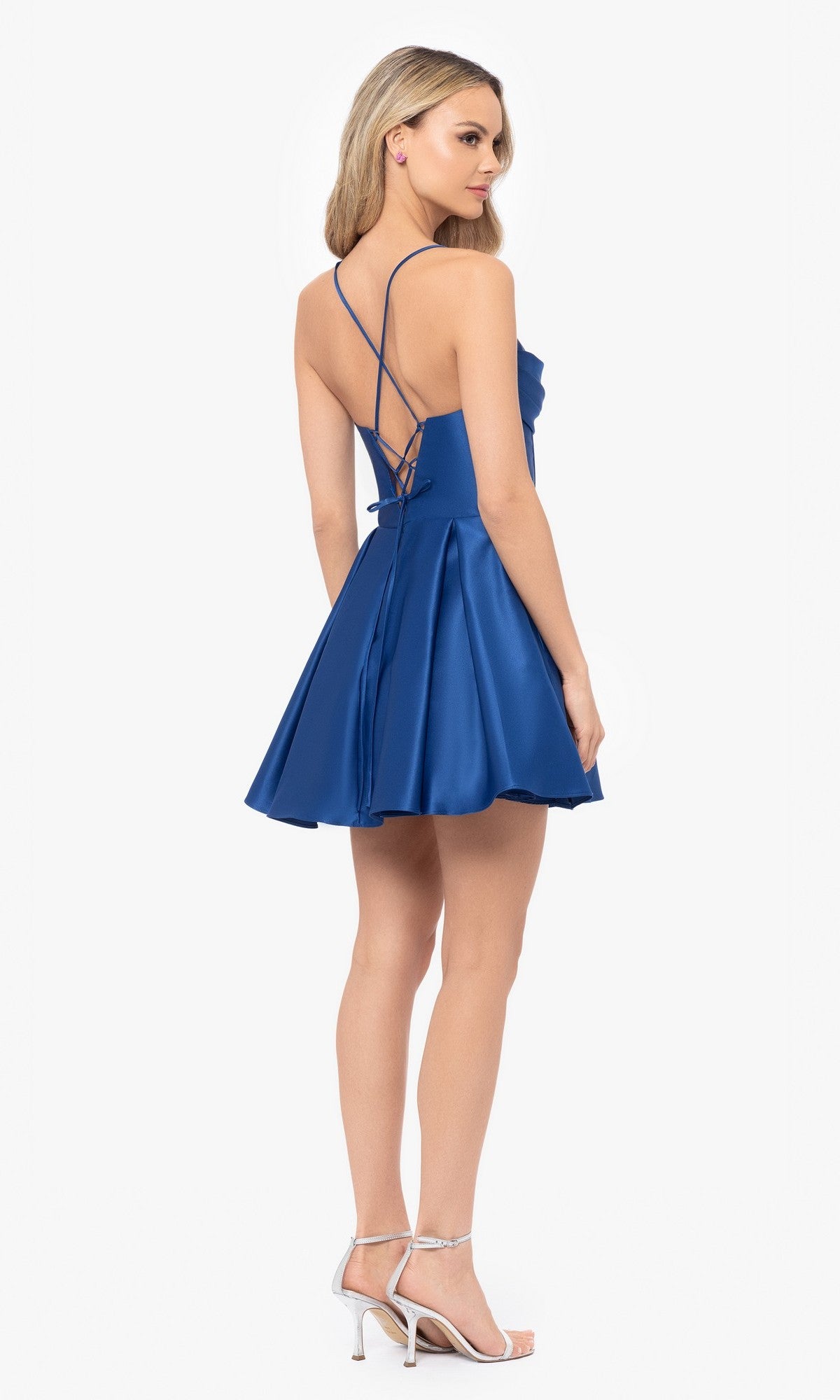A woman wears the Lace-Up Short Hoco Dress: Blondie Nites 4578BN—a sleeveless royal blue A-line prom dress with crisscross straps and open back—paired with silver heels, standing against a white background.